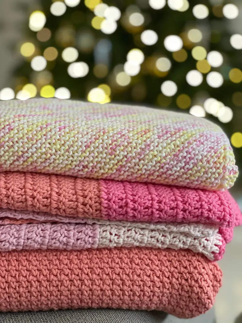 A stack of folded pastel knitted baby blankets in front of blurred holiday lights