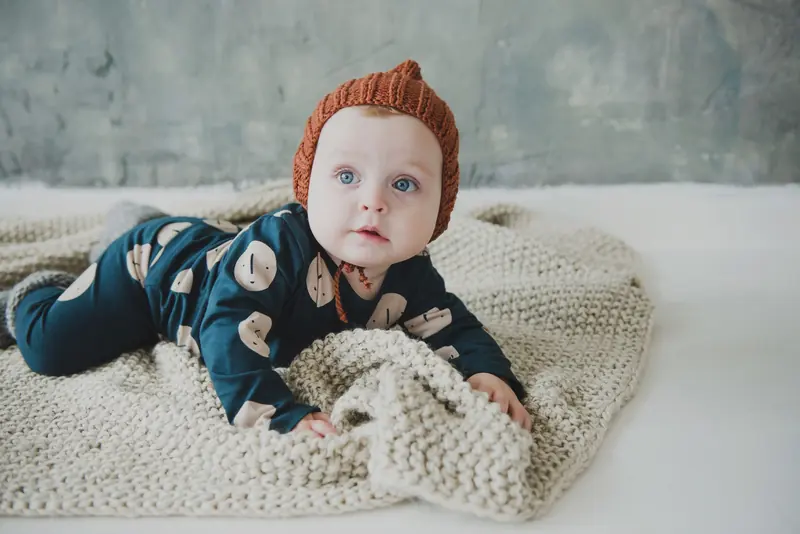 A baby wearing a rust knitted hat with a small top knot, lying on a cream knitted blanket