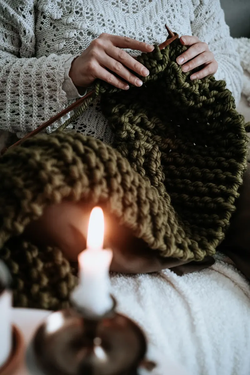 Hands knitting a chunky olive-green throw by candlelight