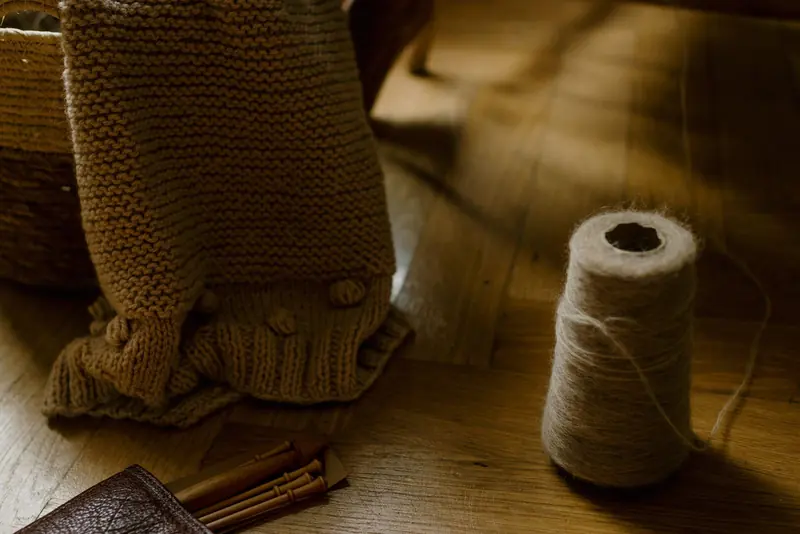 A tan knitted blanket with bobbles draped over a basket beside a cone of yarn