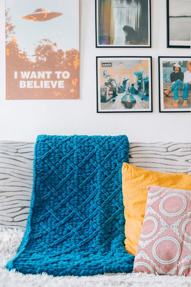 A teal chunky knitted blanket with a diamond texture draped over a sofa