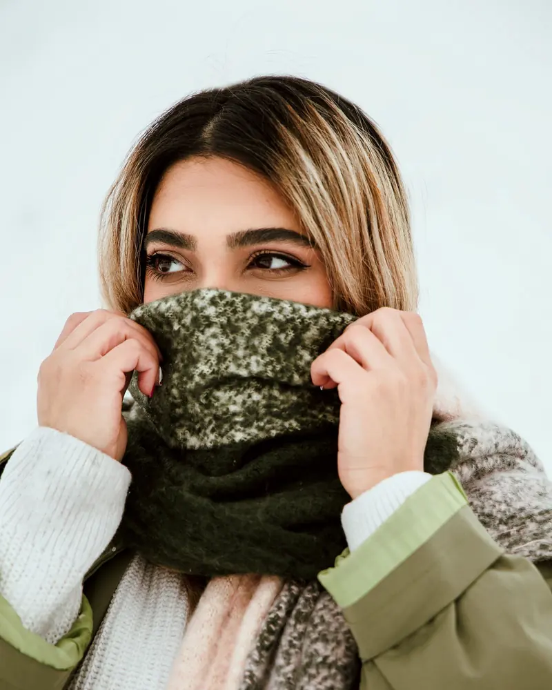 A person holding a speckled green knitted cowl up to their face in winter light