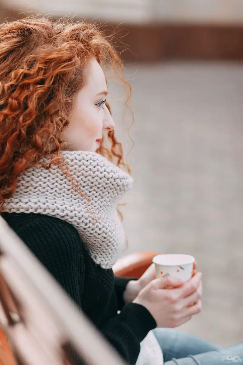 A person wearing a thick beige knitted cowl, holding a cup