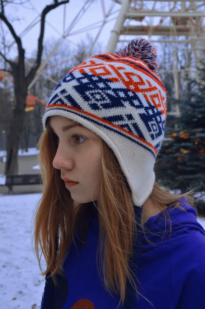 A person wearing a patterned knitted earflap hat with a pom-pom outdoors in snow
