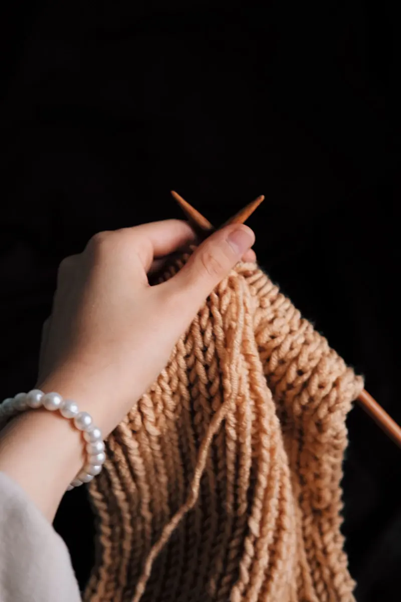 Hands knitting a chunky tan ribbed piece with wooden needles against a dark background