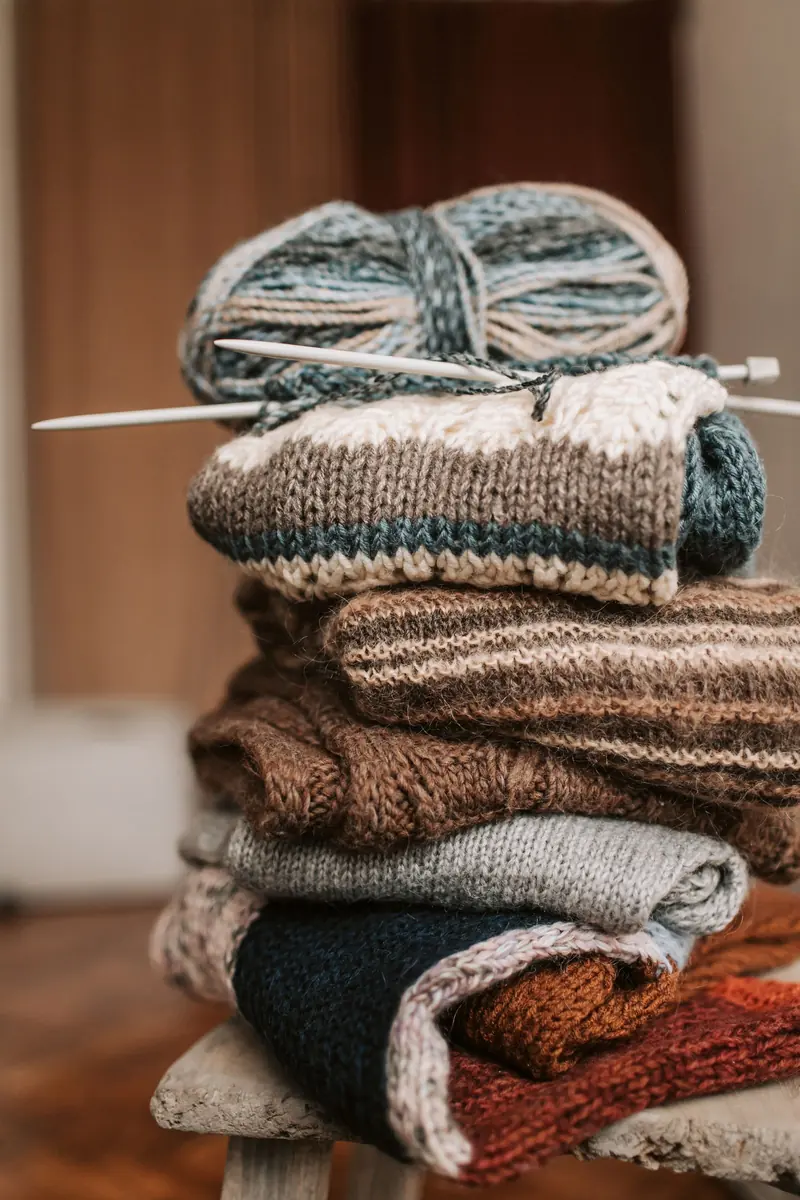 A stack of folded knitted scarves in earthy stripes with a yarn ball and needles on a wooden stool