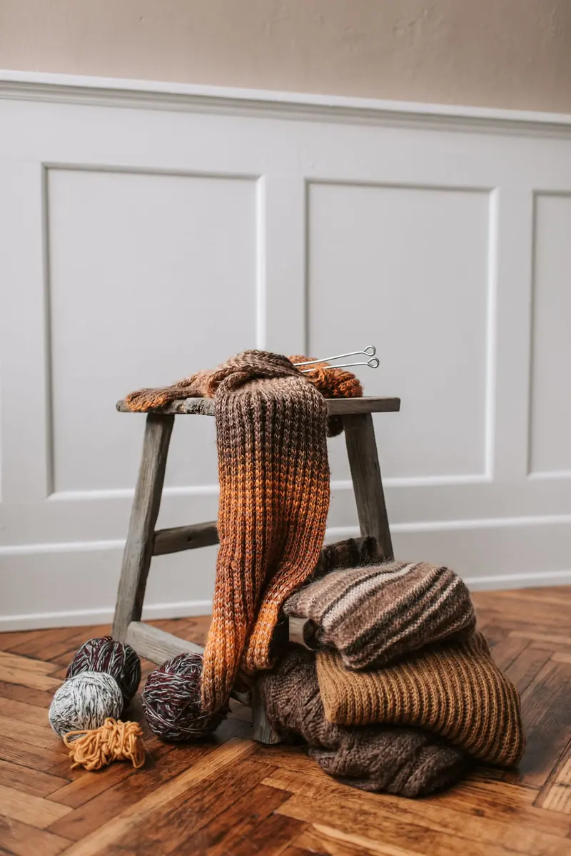 A brown-to-orange ribbed knitted scarf draped over a wooden stool beside yarn balls
