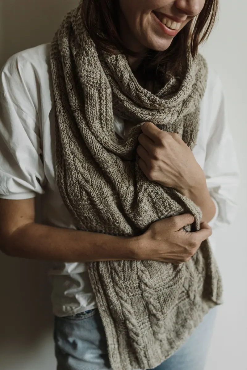 A person holding a thick beige cable-knit neck wrap against a plain wall